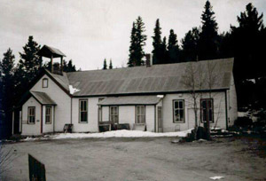 A black and white photo of the building with sloping roof, and extension on the left, in the background are tall evergreen trees. 
