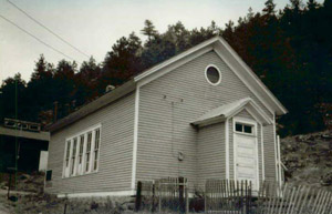 A black and white photo of the building with gabled roof, series of white trimmed windows bunched together on the side and inset small gabled extension on the end which houses the door.