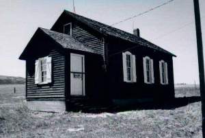 A black and white photo of the dark school house with gabled roof, white shudders and white door on the left. 