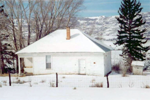 A picture of the school with hipped roof and white walls. Around and on the building is a layer of snow with some trees behind it.