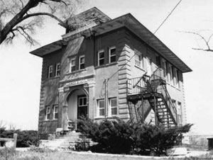 A black and white photo of the building with overhanging roof and smaller floor above. On the right there is a fire escape. 