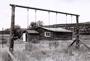 A black and white photo of the house with log walls and wide gabled roof with protruding gabled entrance in front. Before the school stands a a swing set. 
