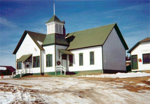 A photo of the school with hip roof tower in the center and cross gables on the right. The roof and siding are green and the siding is white. 