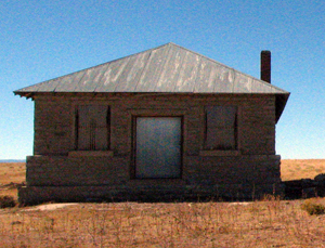 A front view of the building with brick wall with central entrance and two windows on either side with hipped roof and chimney on the right with a field around.