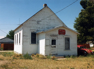 A view of the white school with fading walls and gabled roof before an extended section with one door entrance. 
