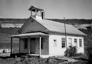 A black and white photo of the school with white walls and gable roof bell tower over the covered entrance. 