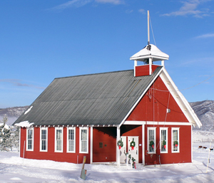 A picture of the brightly painted red school with gable roof and white trim. In the center near the entrance covered in the corner stands a red bell tower. The school is surrounded by snow and you can see some mountains in the background.