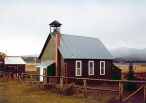 A photo of the school with green walls, pitched metal roof and bell tower on top. In the background are distant mountains and before the school stands a short log fence.