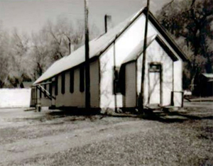 A black and white photo of the school with white walls and overhanging gable roof and smaller protruding entrance. 